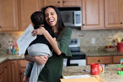 Adult embracing a child in a kitchen, both smiling, with a diploma in hand and various household items like a mug and papers on the counter.